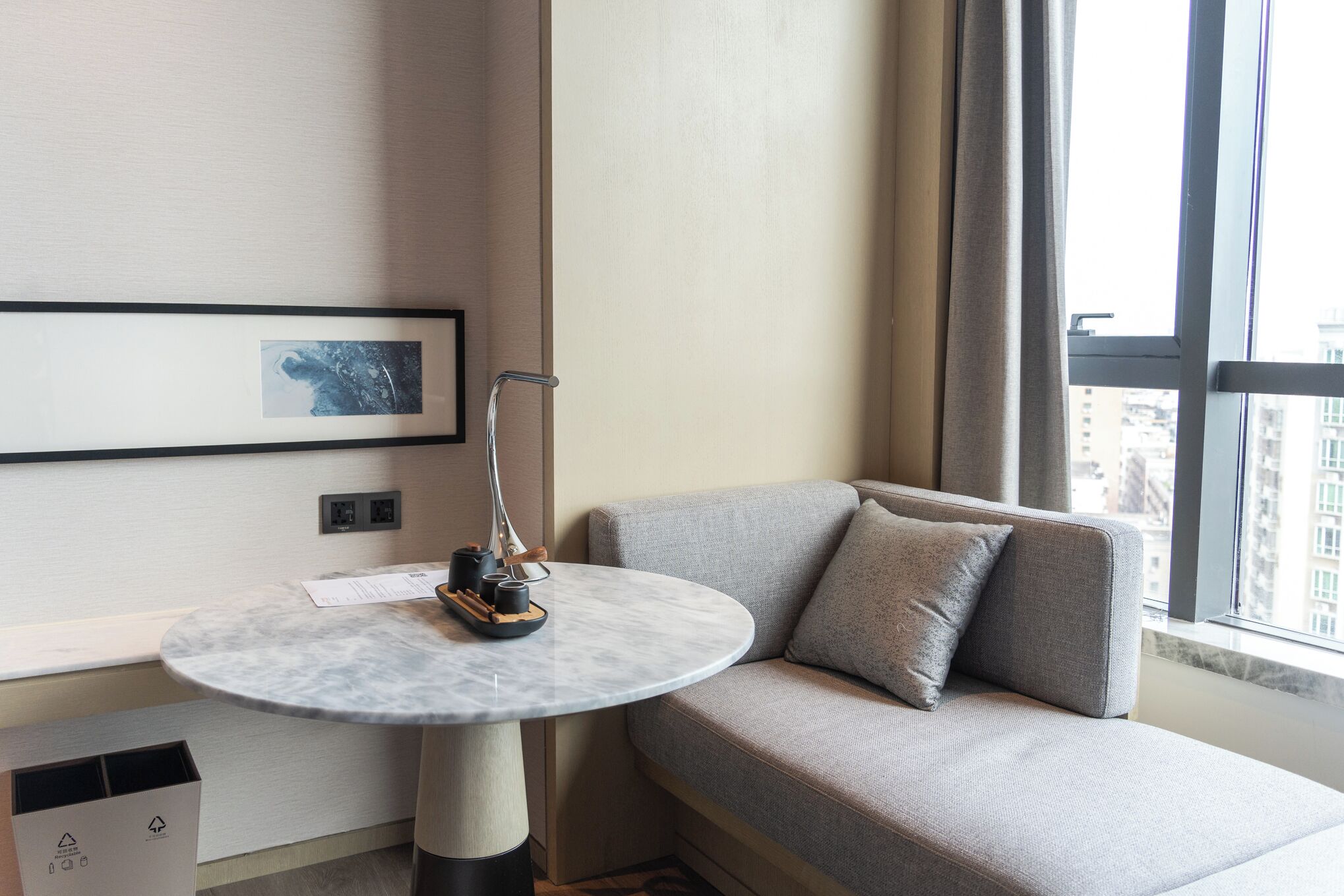 A modern hotel room with a round marble table, a grey sofa, and a city view through large windows.