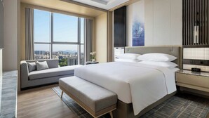 Une chambre d’hôtel moderne avec un grand lit, un canapé et un banc. Il y a une fenêtre donnant sur la ville.
