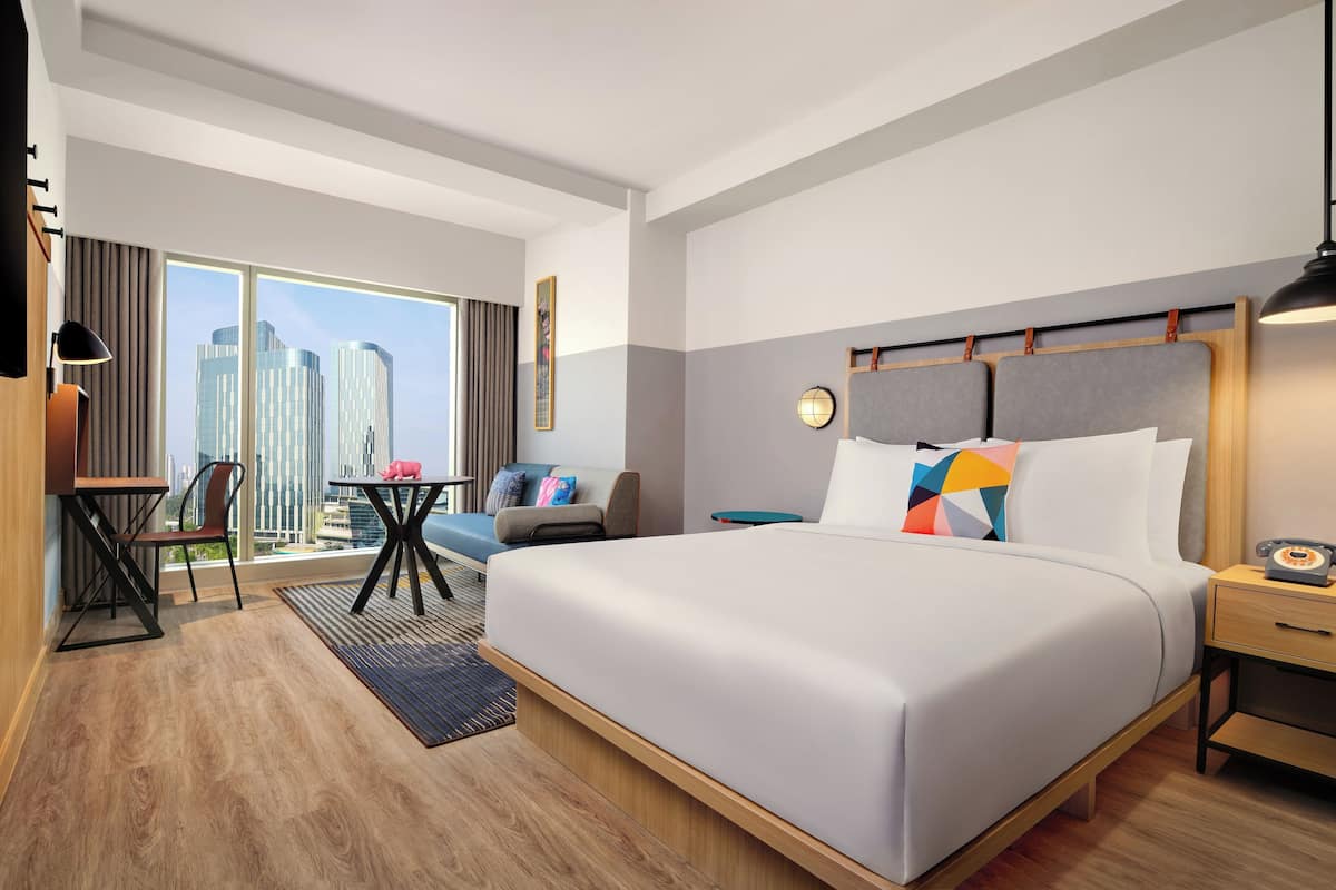 A modern hotel room with a large bed, a desk, a chair, and a view of the city skyline.