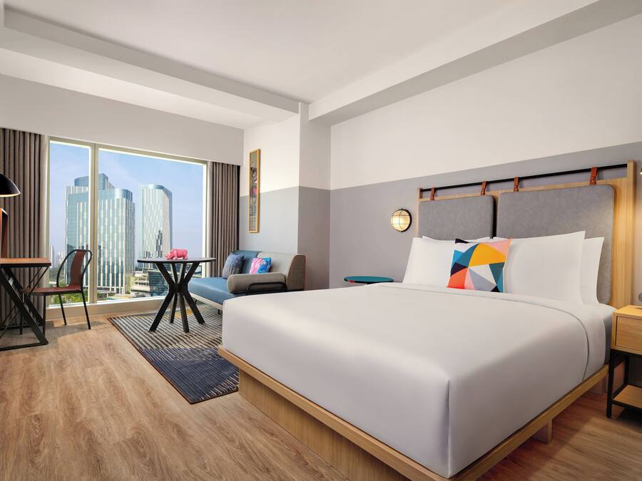 A modern hotel room with a large bed, a desk, a chair, and a view of the city skyline.