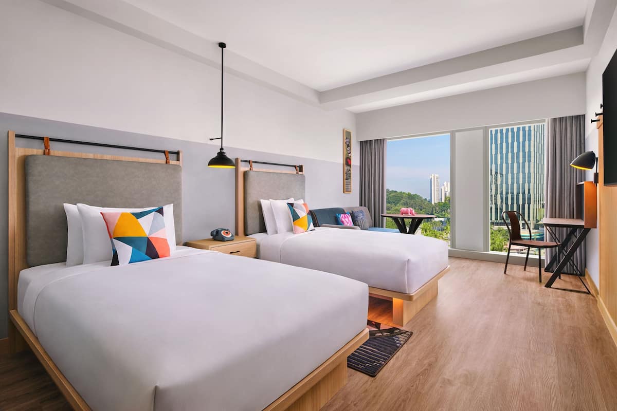 A hotel room with two beds, a desk, and a chair. There is a large window with a view of buildings and greenery.