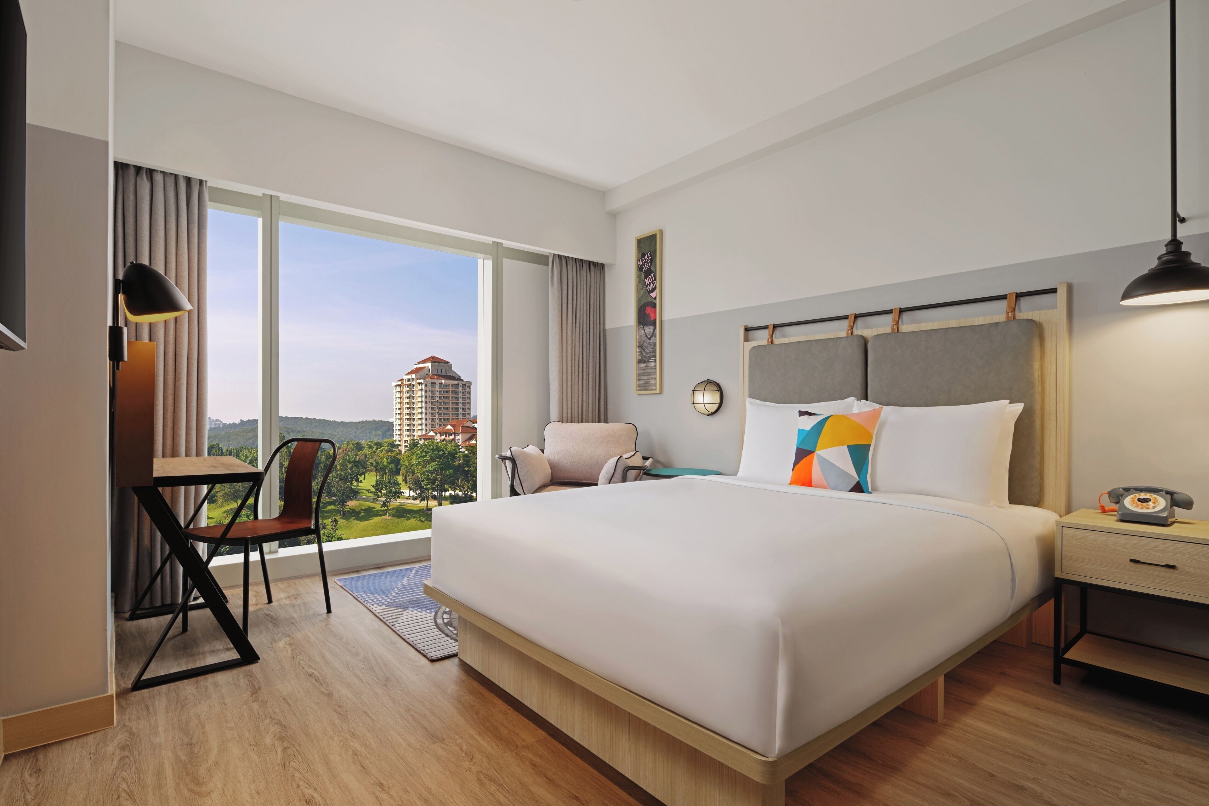 A hotel room with a large bed, a desk with a chair, a nightstand, and a view of buildings through large windows.