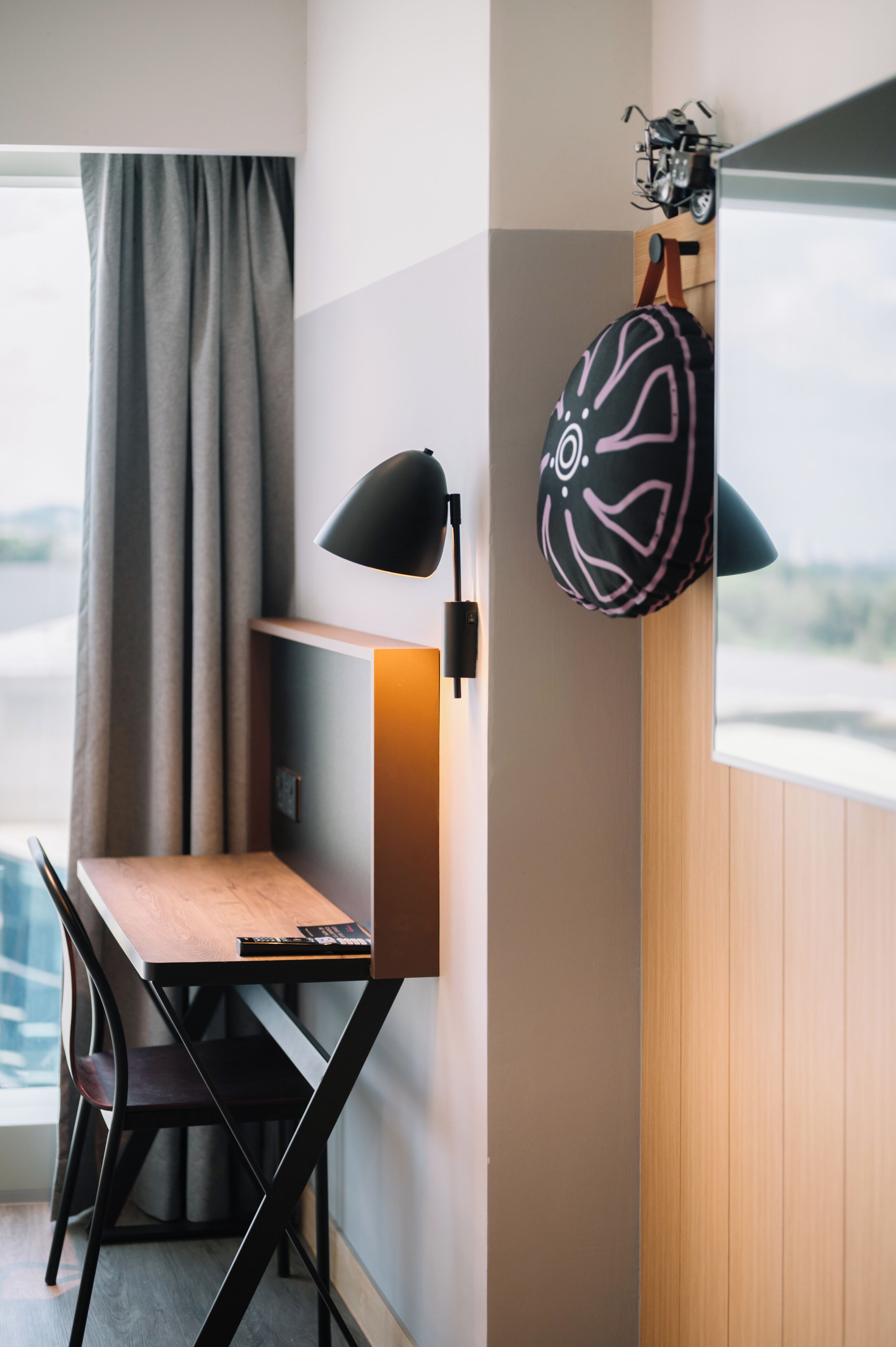 A compact workspace with a desk, chair, and lamp, a window with curtains, and a wall-mounted bag.