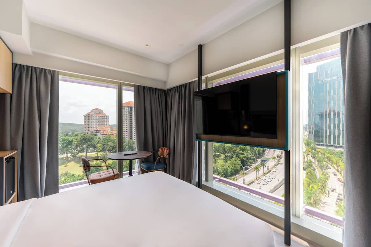 A hotel room with a large bed, a TV mounted on the wall, a small table with a chair, and a view of a cityscape through large windows.