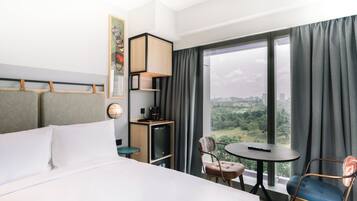 A hotel room with a bed, a desk, a chair, a small table, a TV, and a large window with a view of a city and greenery.