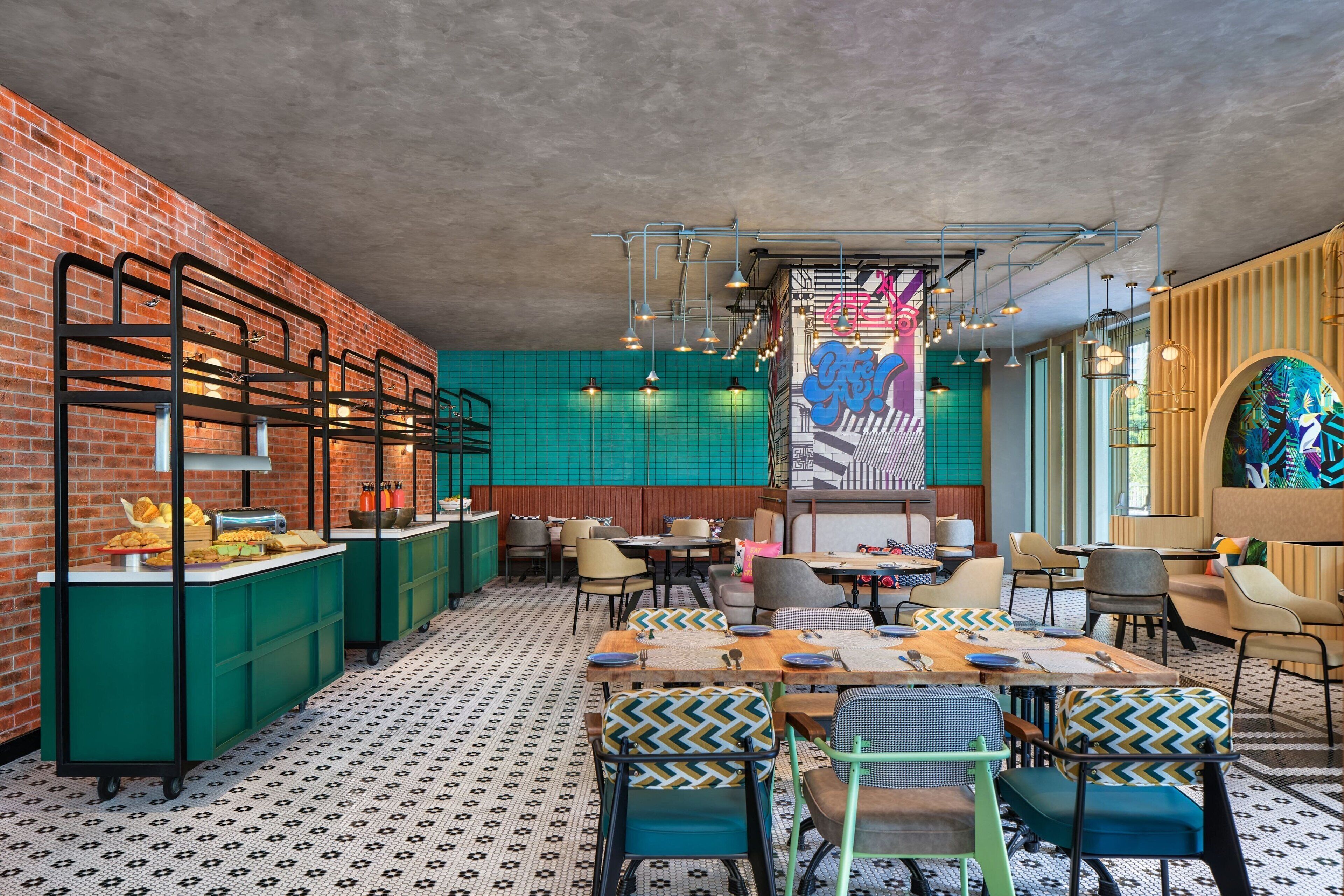 Ein Restaurant mit Backsteinwand, türkis schimmernden Fliesen und einem gemusterten Boden. Tische sind mit Stühlen und Tischdekorationen gedeckt.