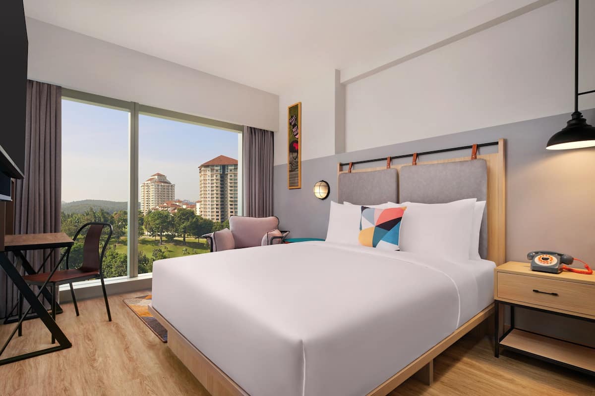A modern hotel room with a large bed, a desk, and a chair. There is a view of buildings and greenery outside the window.