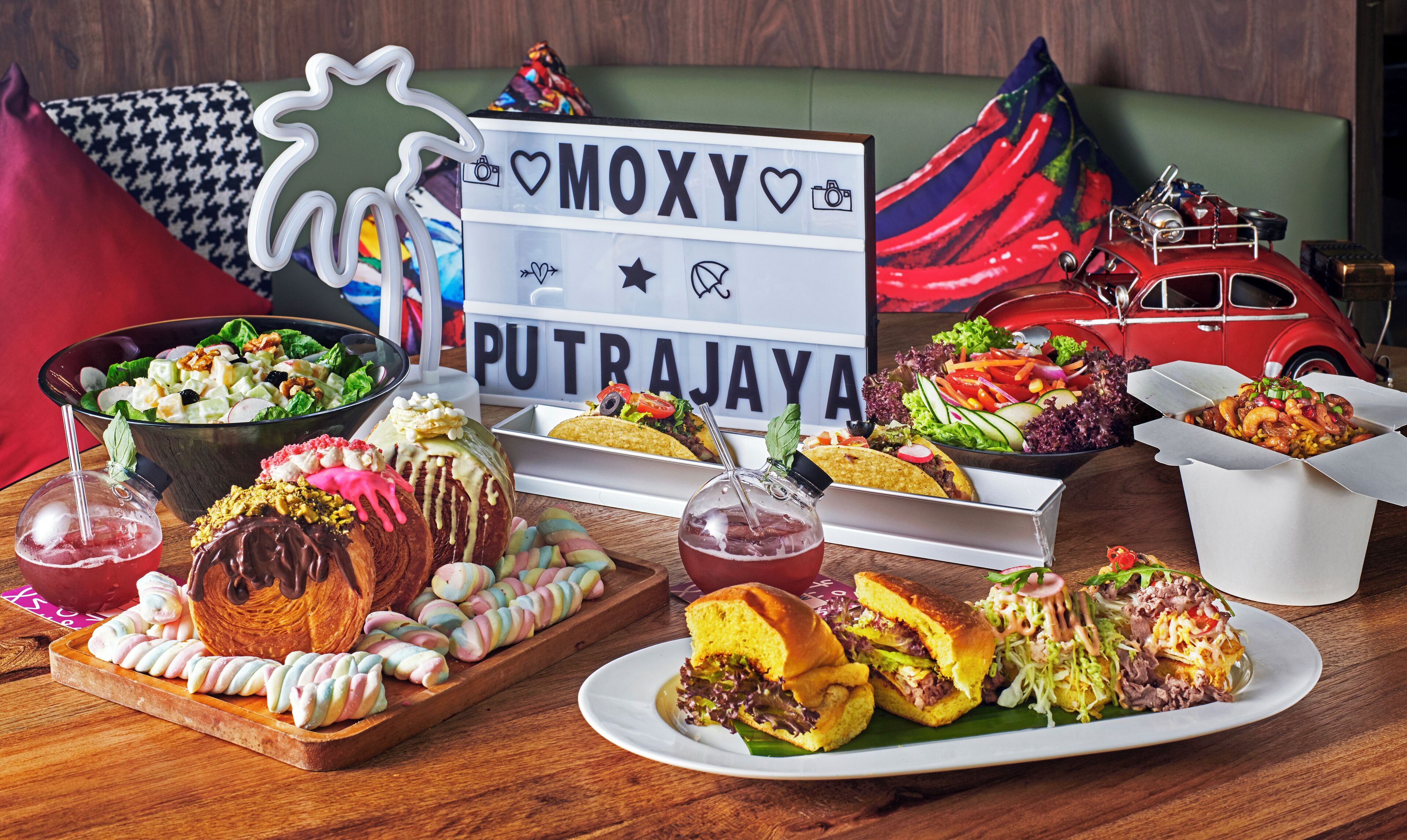 A variety of food items including a sandwich, tacos, and a salad, displayed on a wooden table with a vintage car and a sign reading "MOXY PUTRAJAYA".