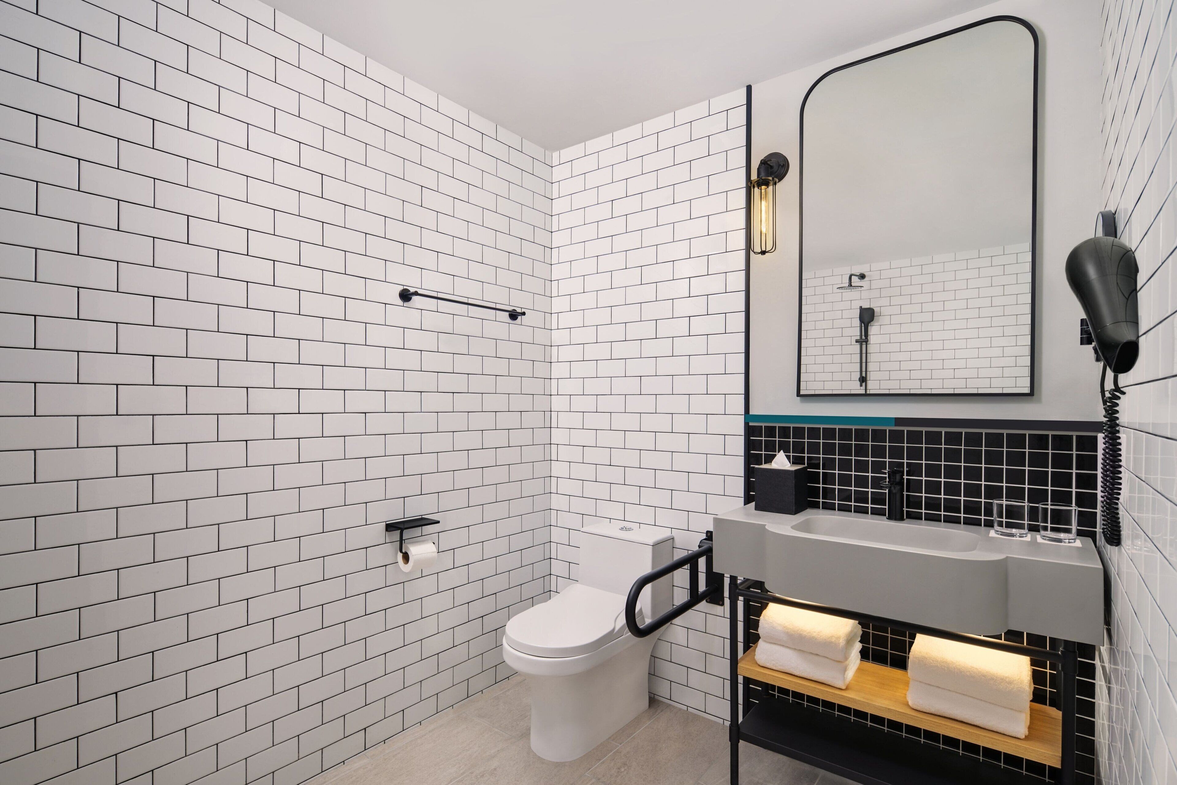 A modern bathroom with white subway tiles, a white toilet, a black towel rack, a black hairdryer, and a mirror with a black frame.