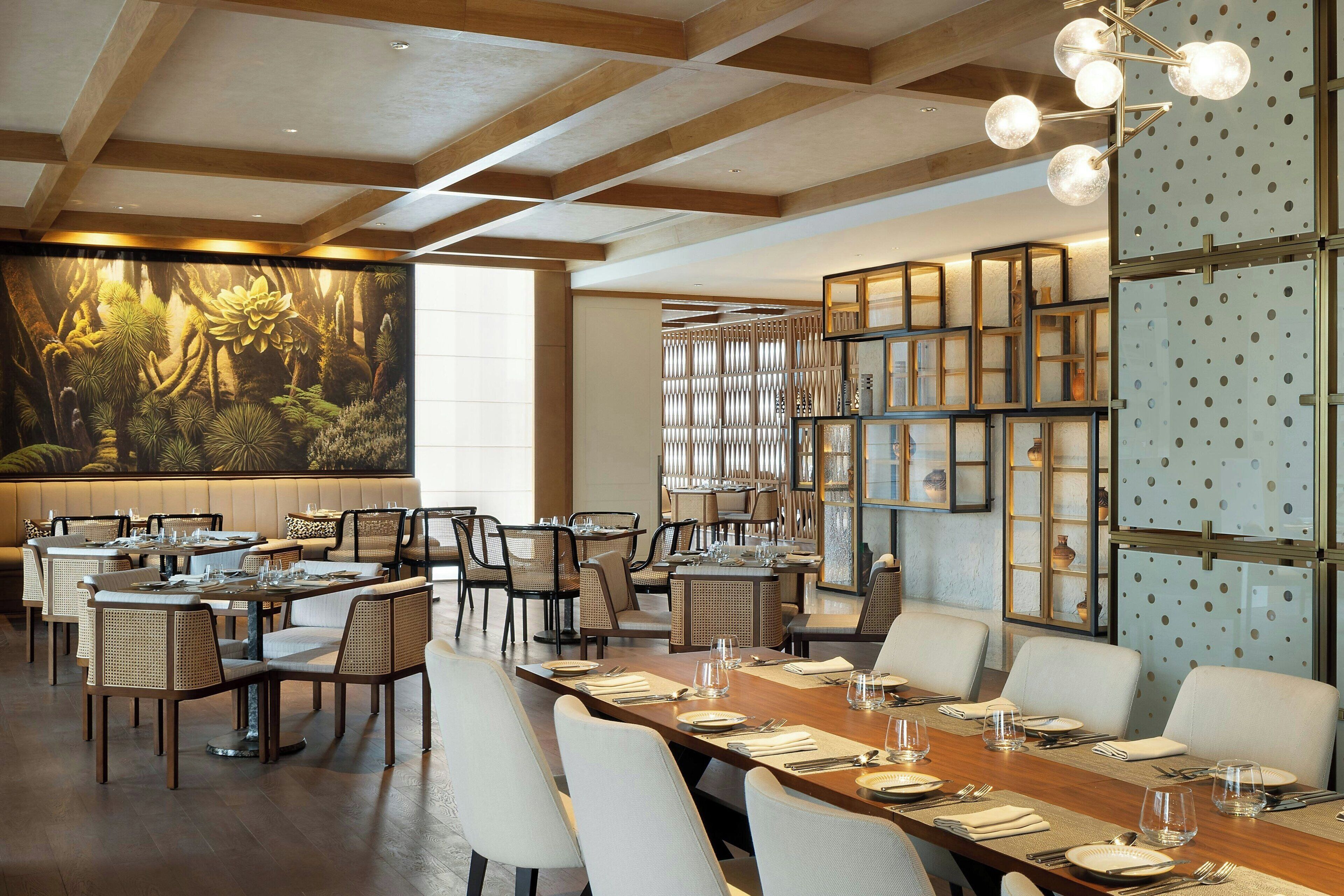 A spacious dining area with high ceilings and a large mural of a forest scene. Tables are set with white tablecloths and chairs, and there are glass display cases with decorative items.