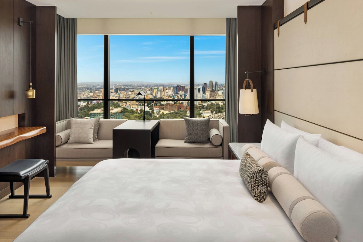 A hotel room with a large bed, a sofa, a small table, and a view of a cityscape through large windows.