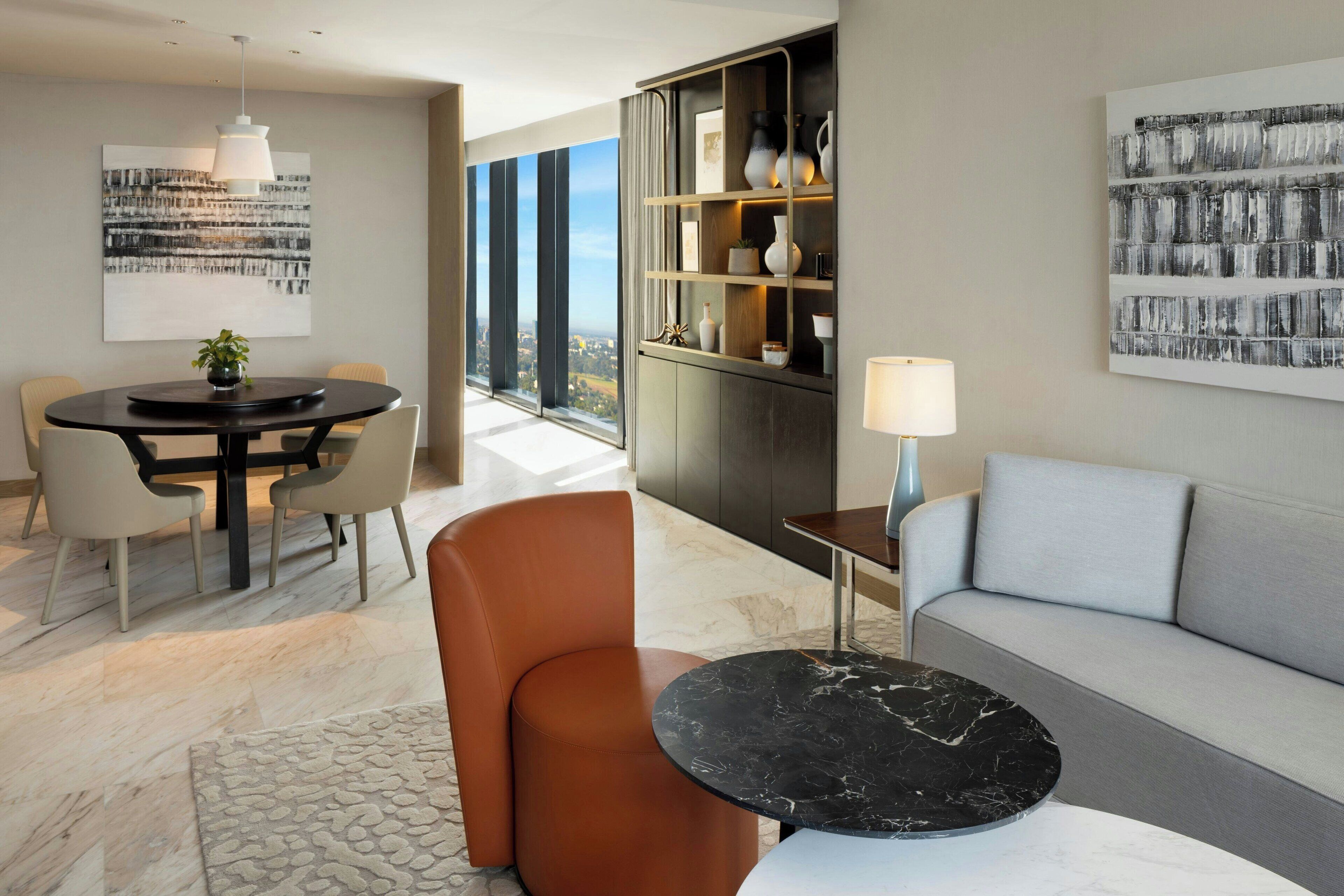 A modern living room with a round dining table, a sofa, and a coffee table. There is a large window providing a view of the city.