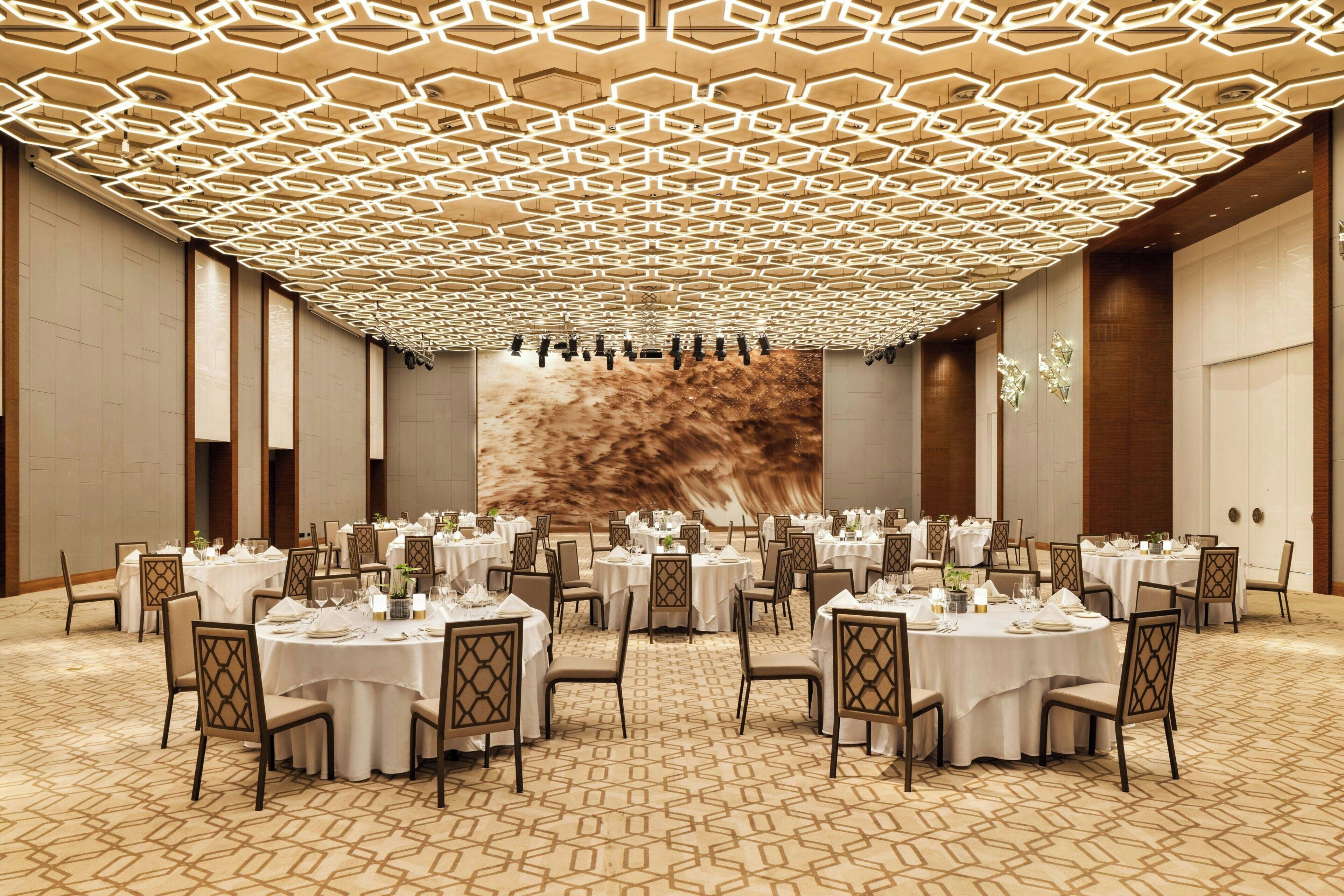 A banquet hall with round tables set for dining, a patterned carpet, and a geometric ceiling design.