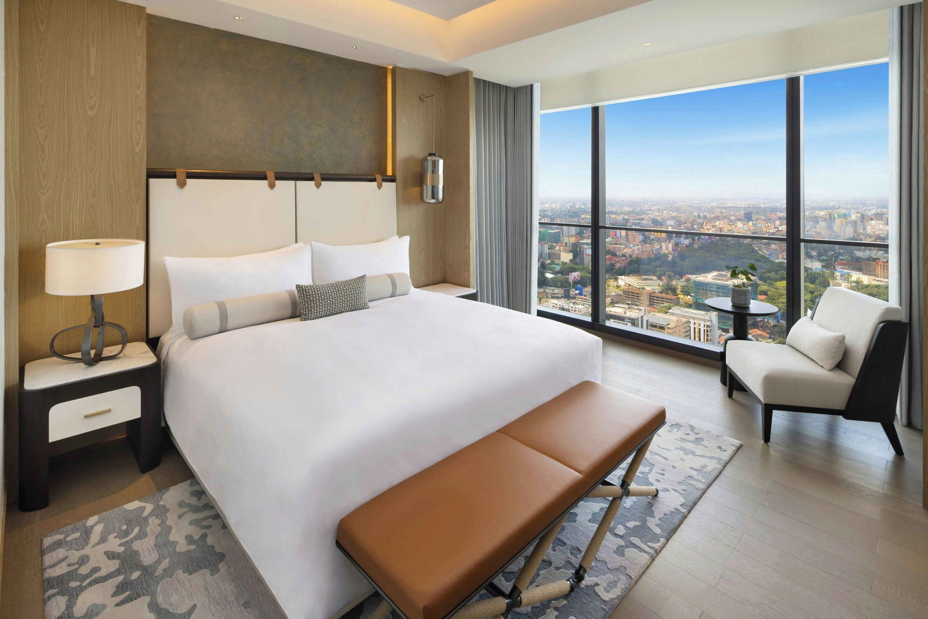 A modern hotel room with a large bed, a bench, a chair, a nightstand, and a view of the city through large windows.