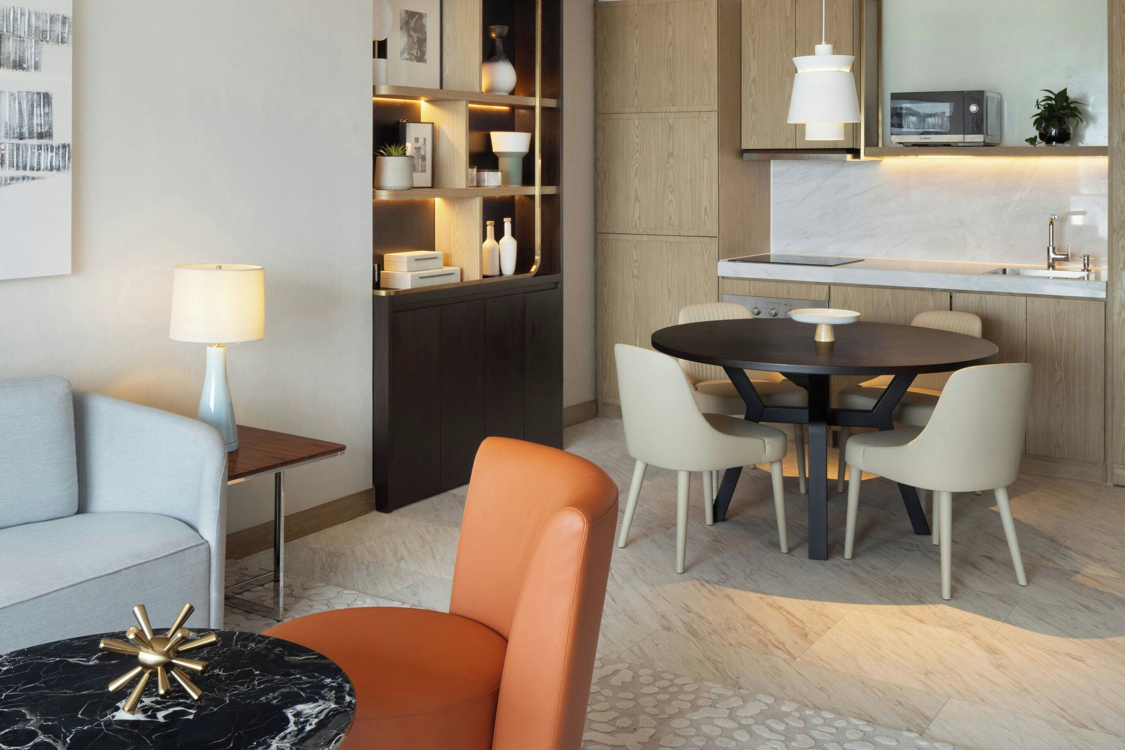 A modern living room with a sofa, a round table with chairs, a coffee table, a lamp, a bookshelf, and a kitchen area with a countertop and cabinets.