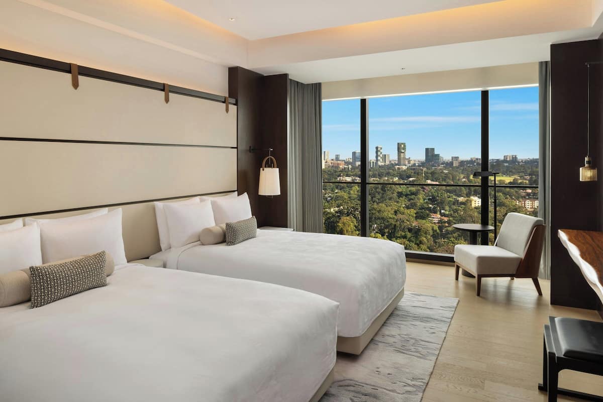 Ein modernes Hotelzimmer mit zwei grossen Betten, Blick auf die Stadt und einem Balkon mit einem Sessel.