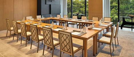 A conference room with a long table, chairs, and a view of a forest through large windows.