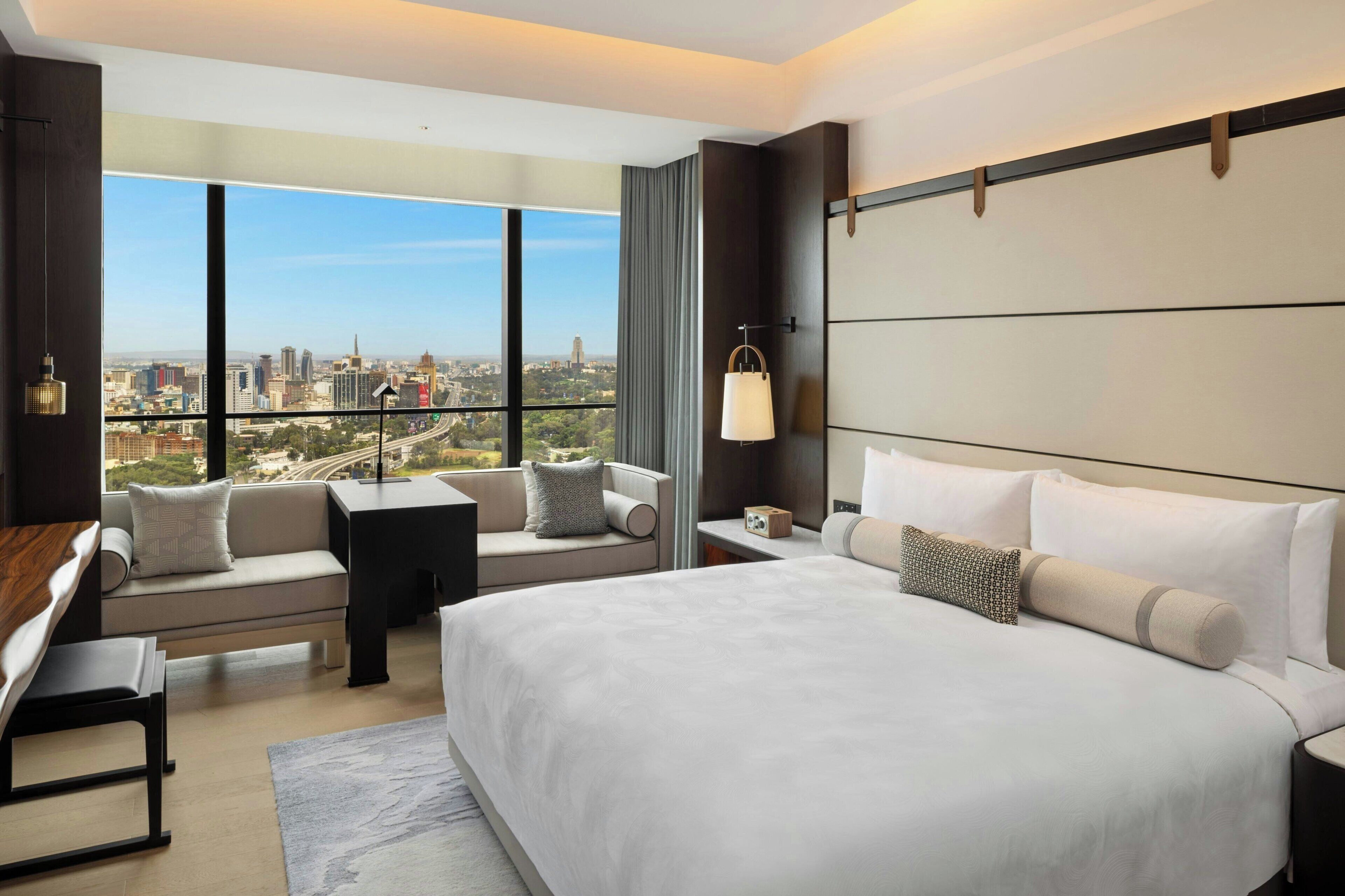Une chambre d’hôtel moderne dotée d’un grand lit, d’un canapé, d’un bureau et offrant une vue sur le paysage urbain grâce à de grandes fenêtres.