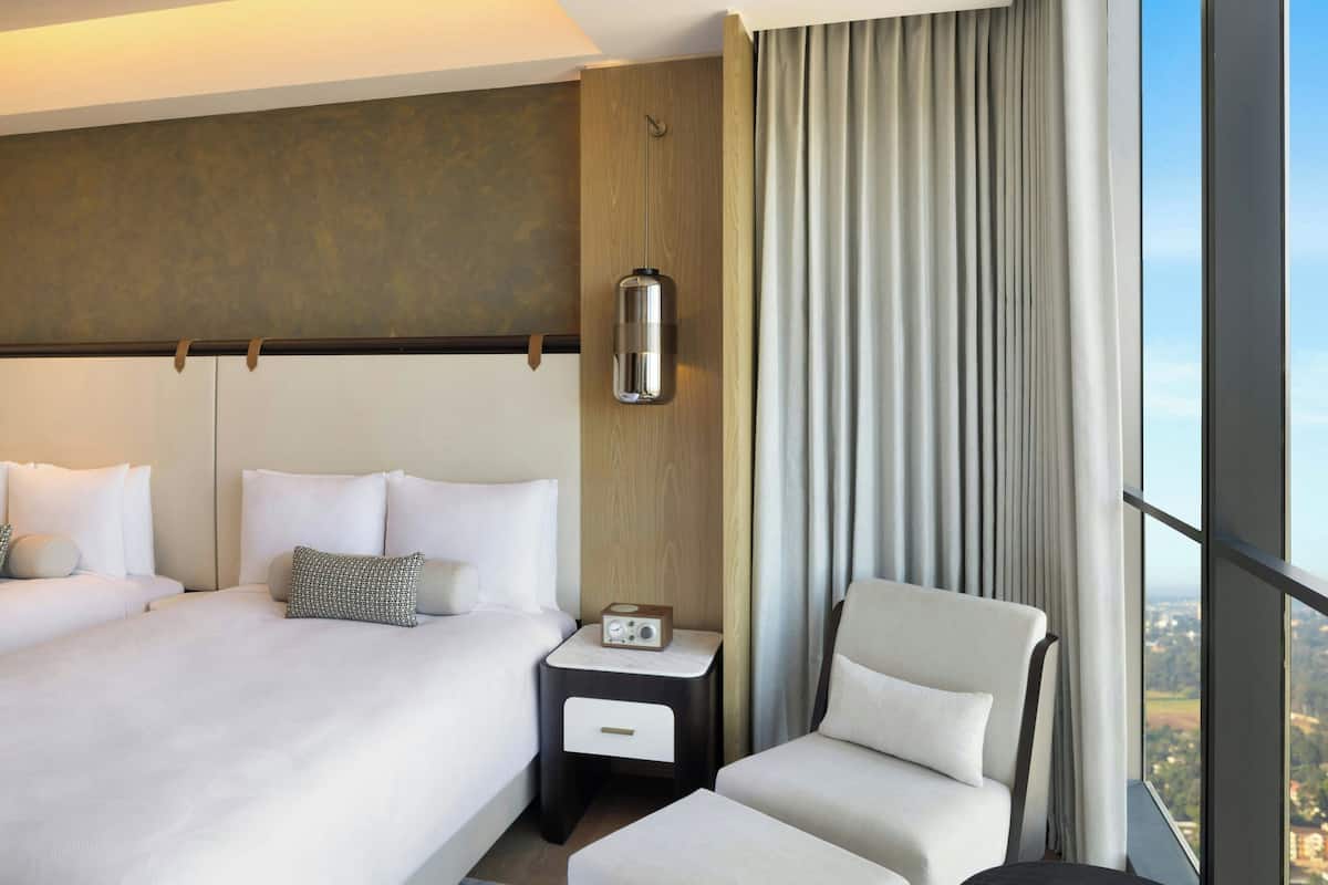 Ein Hotelzimmer mit einem großen Bett, einem Sessel, einem Nachttisch mit Radio und Blick auf die Stadt durch große Fenster.