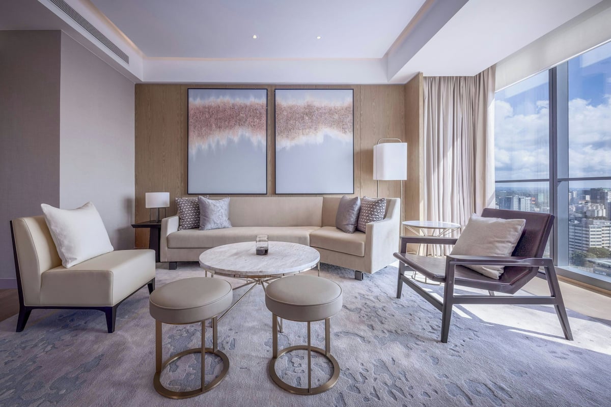 Ein modernes Hotelzimmer mit einer Couch, zwei Sesseln, einem Couchtisch und Blick auf die Stadt durch große Fenster.