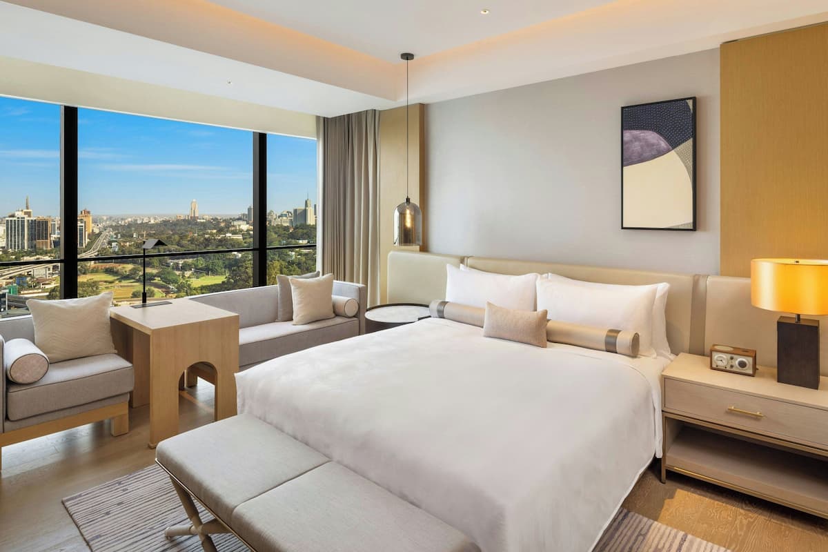 Ein modernes Hotelzimmer mit einem großen Bett, einer Couch, einem kleinen Tisch und einem Nachttisch. Das Zimmer verfügt über ein großes Fenster mit Blick auf die Stadt.