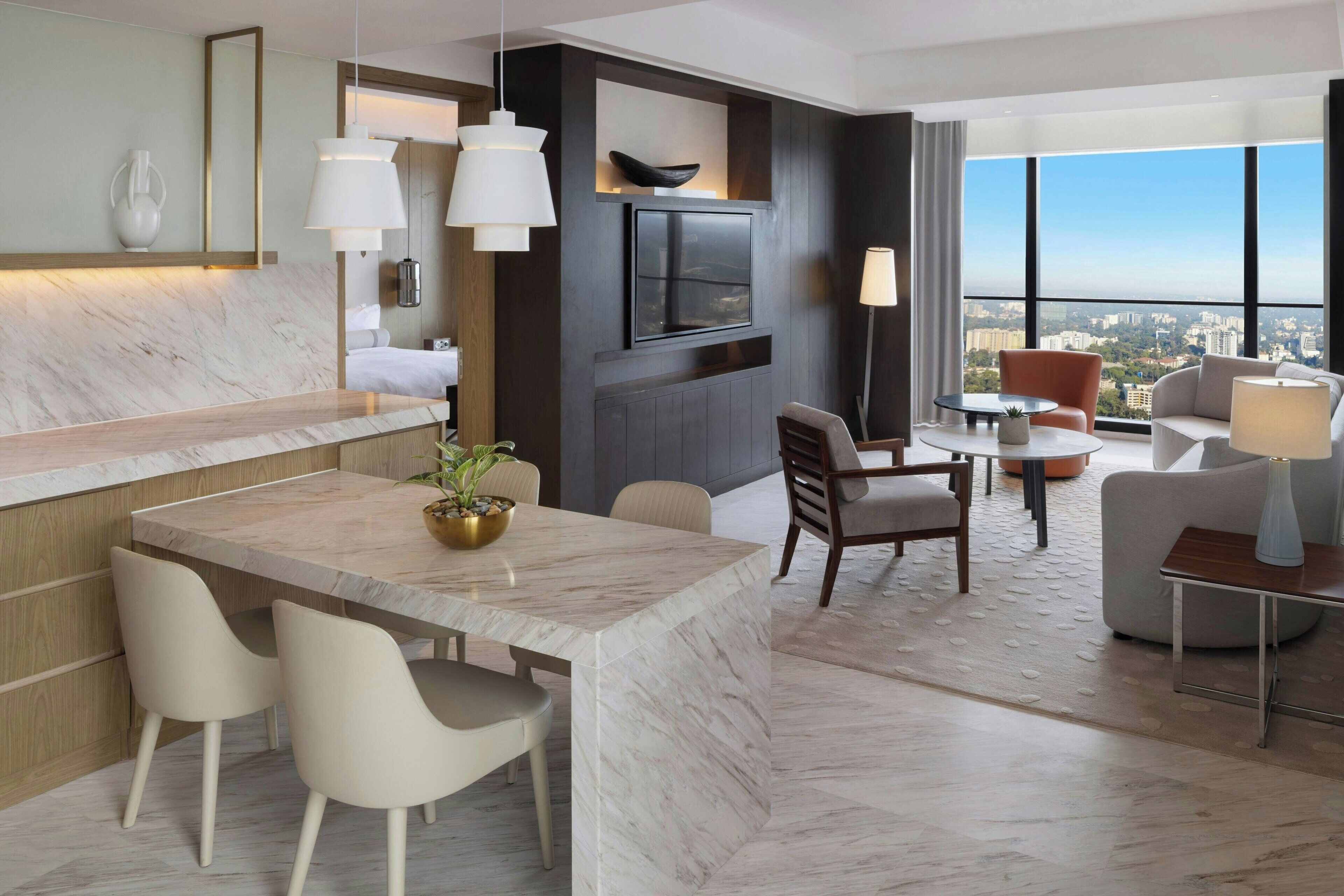 A modern hotel room with a marble dining table, white chairs, a TV mounted on the wall, and a city view through large windows.