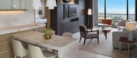 Una habitación de hotel moderna con una mesa de comedor de mármol, sillas blancas, un televisor montado en la pared y vistas a la ciudad a través de amplios ventanales.