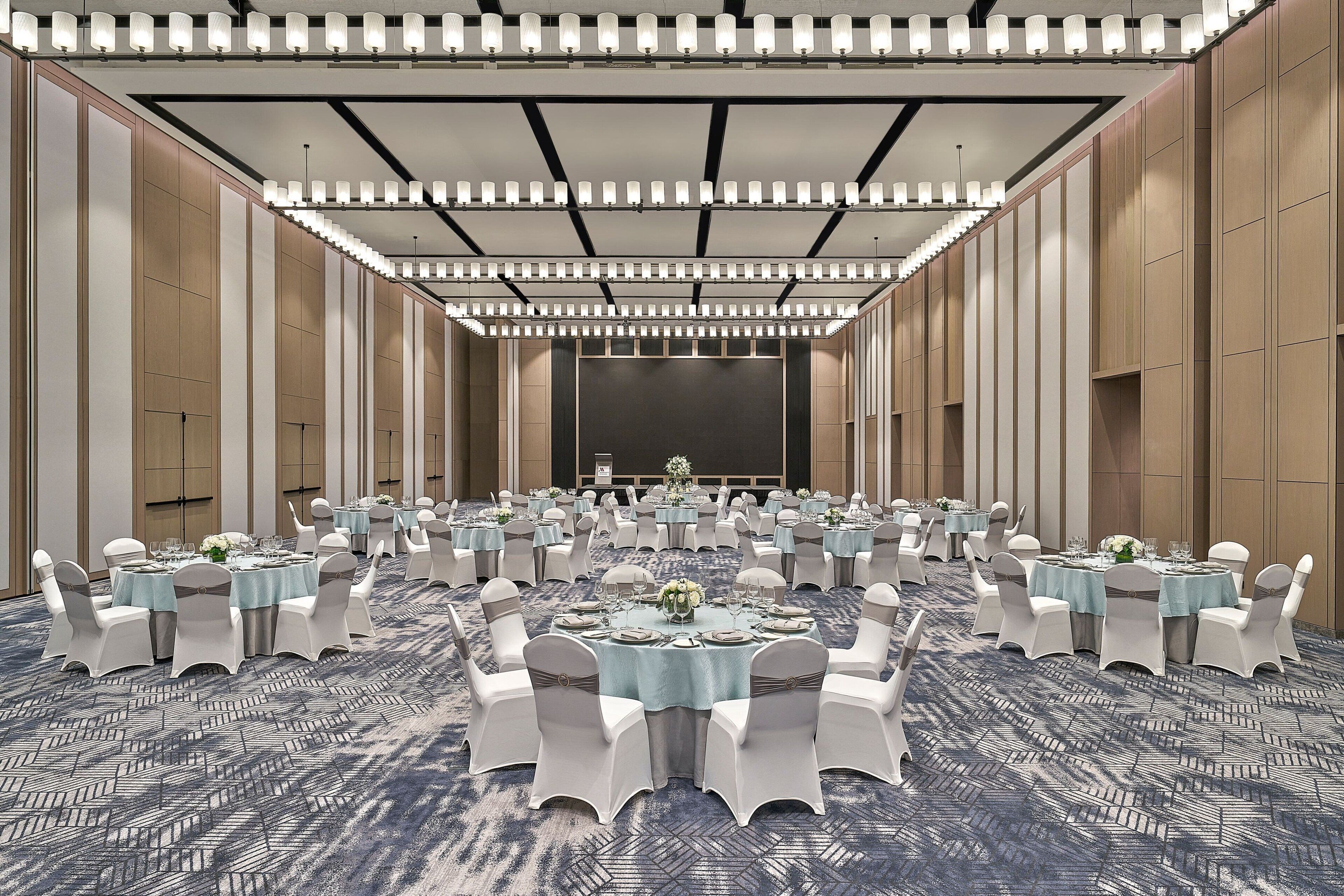 A large banquet hall with multiple round tables, each set with white chairs and blue tablecloths, arranged in rows.
