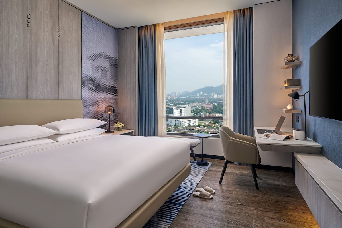 A modern hotel room with a large bed, a desk with a laptop, and a view of a cityscape through a large window.
