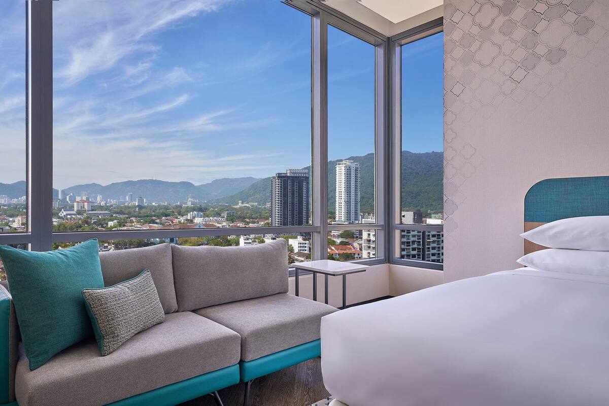 A hotel room with a large window, a sofa with teal and gray cushions, a small table, and a bed with white bedding.