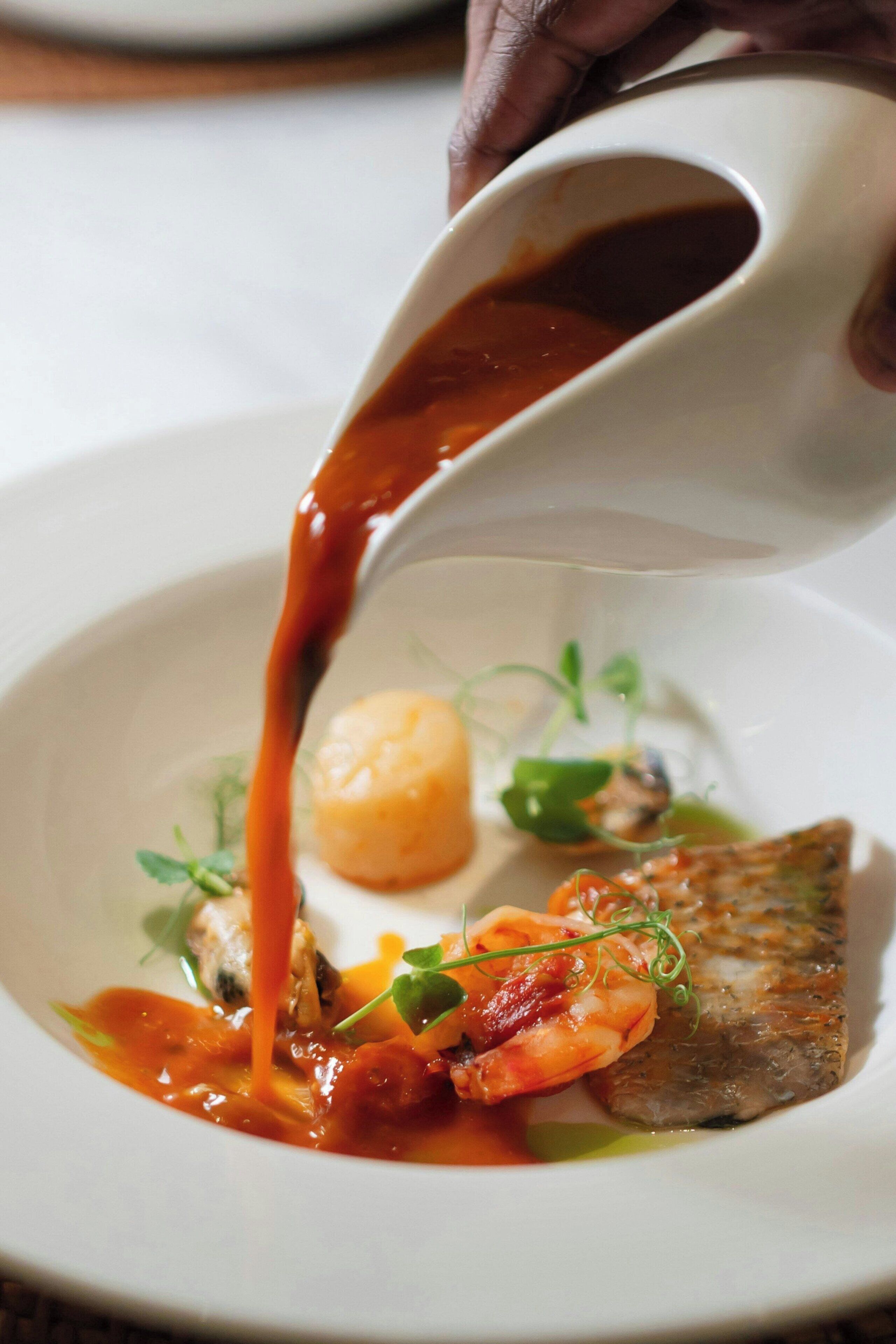 A person pouring sauce over a dish with seafood and greens.