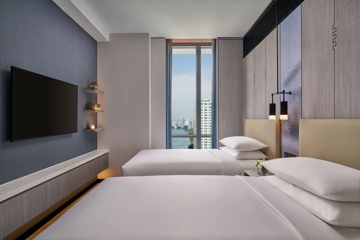 A modern hotel room with a large bed, a TV mounted on the wall, and a city view through the window.