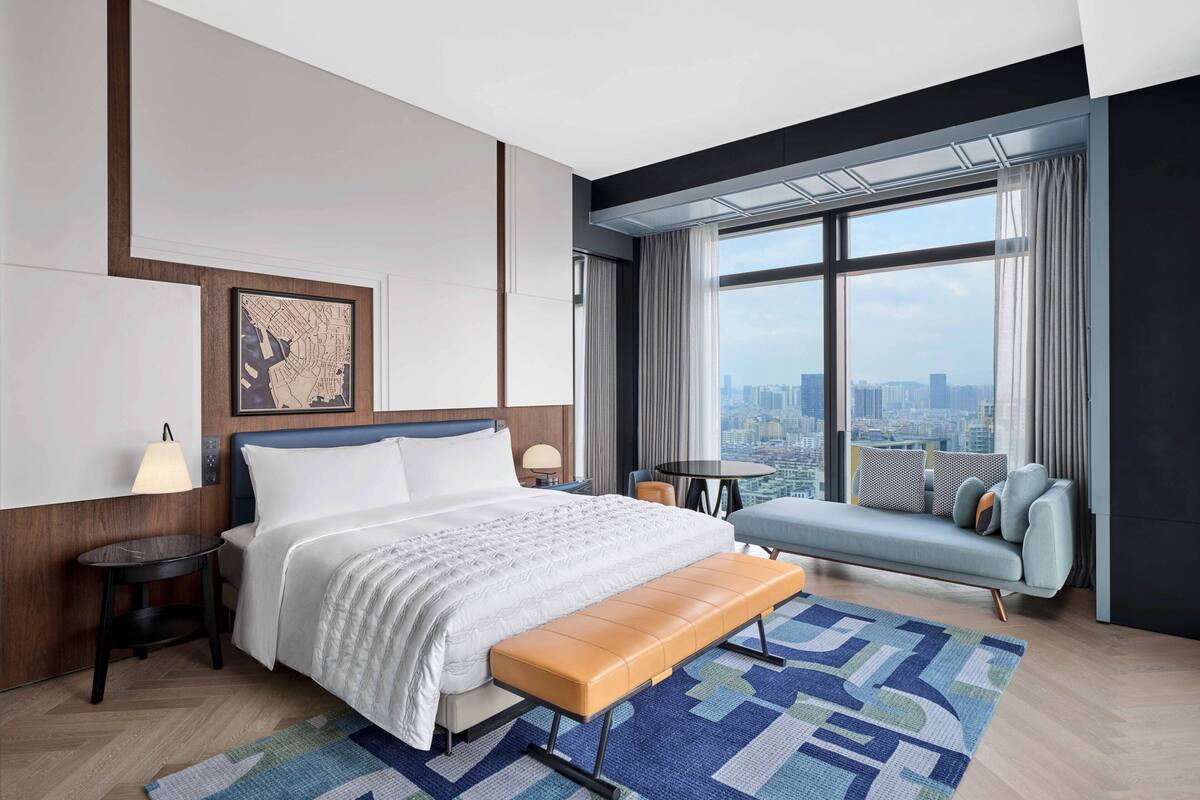 A modern hotel room with a large bed, a bench, a sofa, a city view, and a map on the wall.