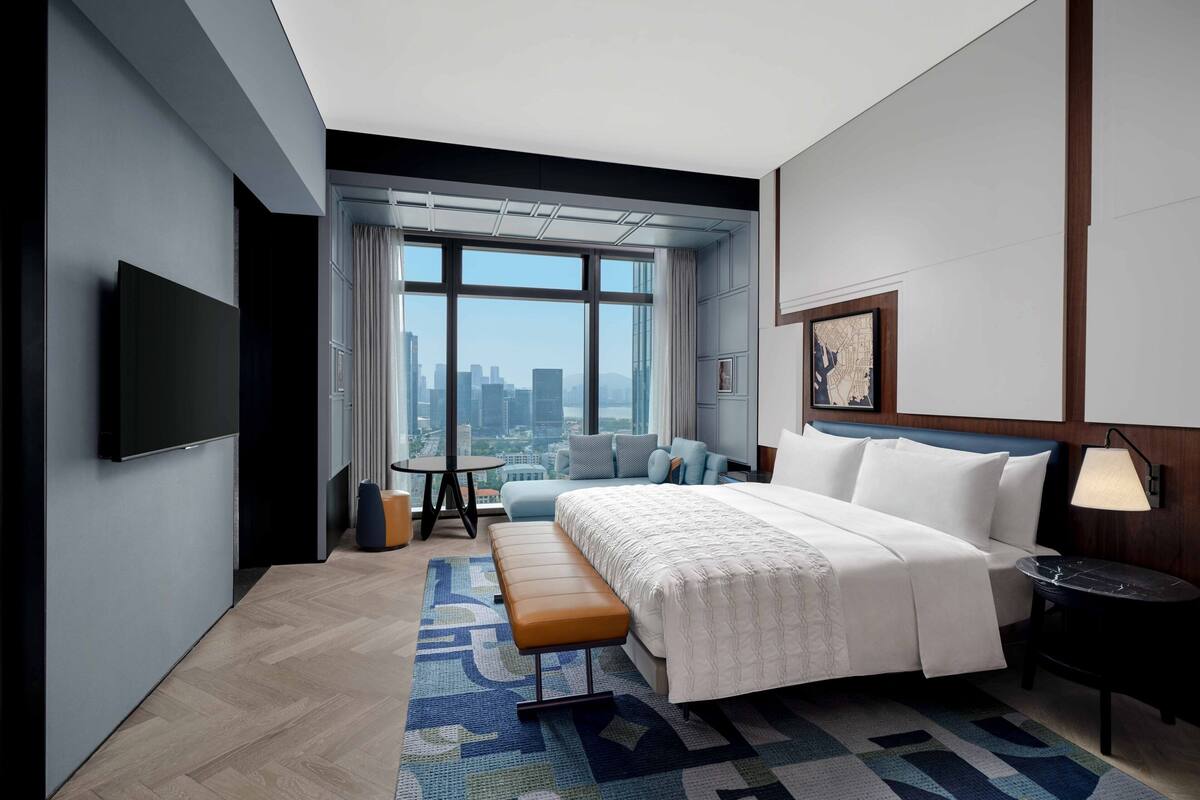 A modern hotel room with a large bed, a seating area, a television, and a city view.