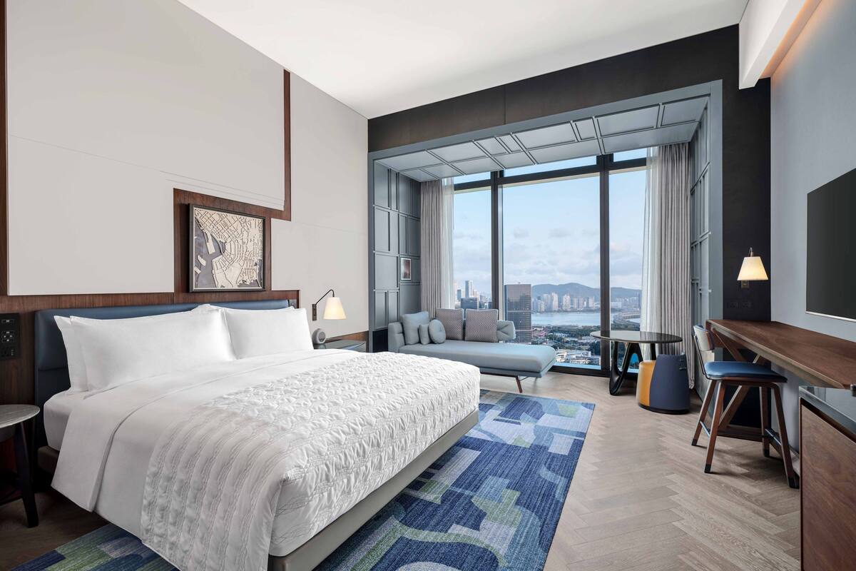 A modern hotel room with a large bed, a sofa, a desk, and a view of the city.