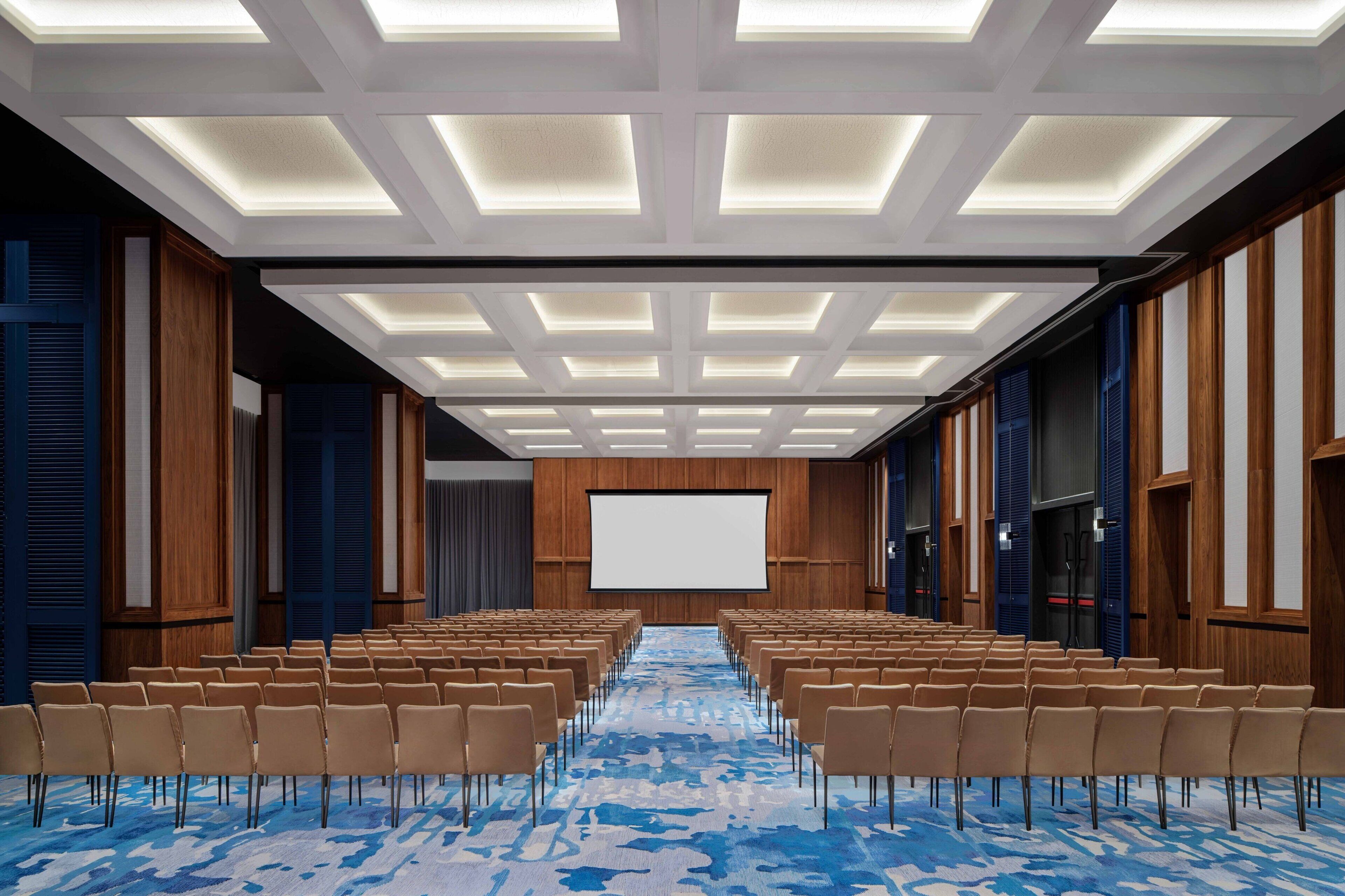 Une salle de conférence avec des rangées de chaises, un grand écran et une moquette à motifs.