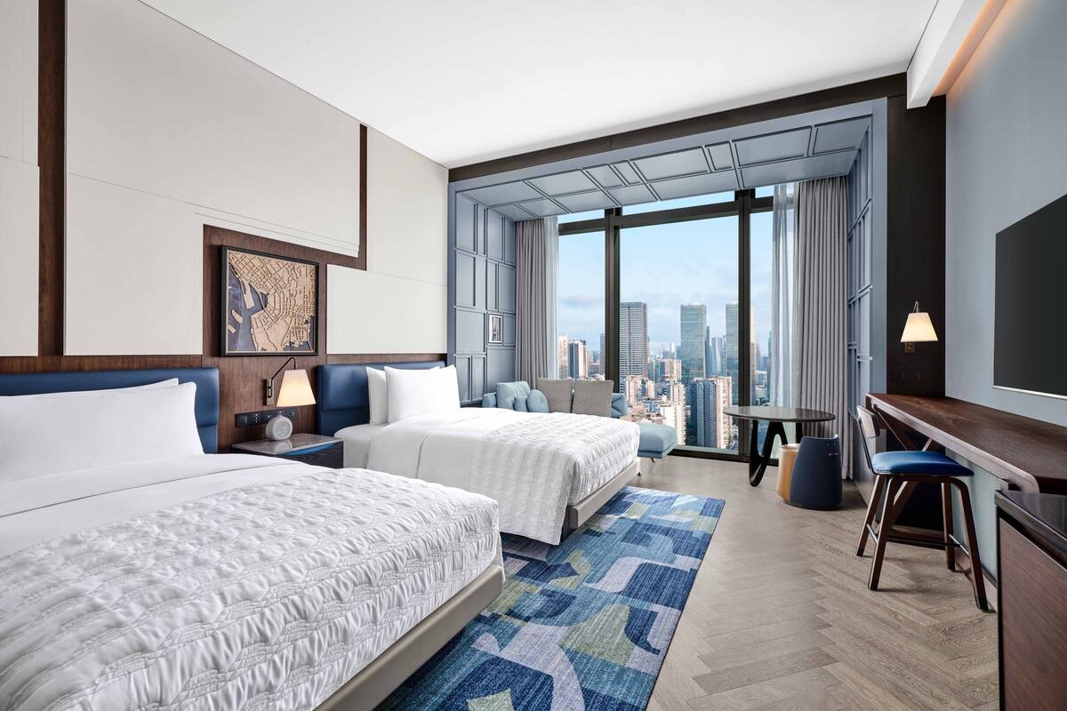 A hotel room with two beds, a desk, a chair, a TV, and a large window with a city view.