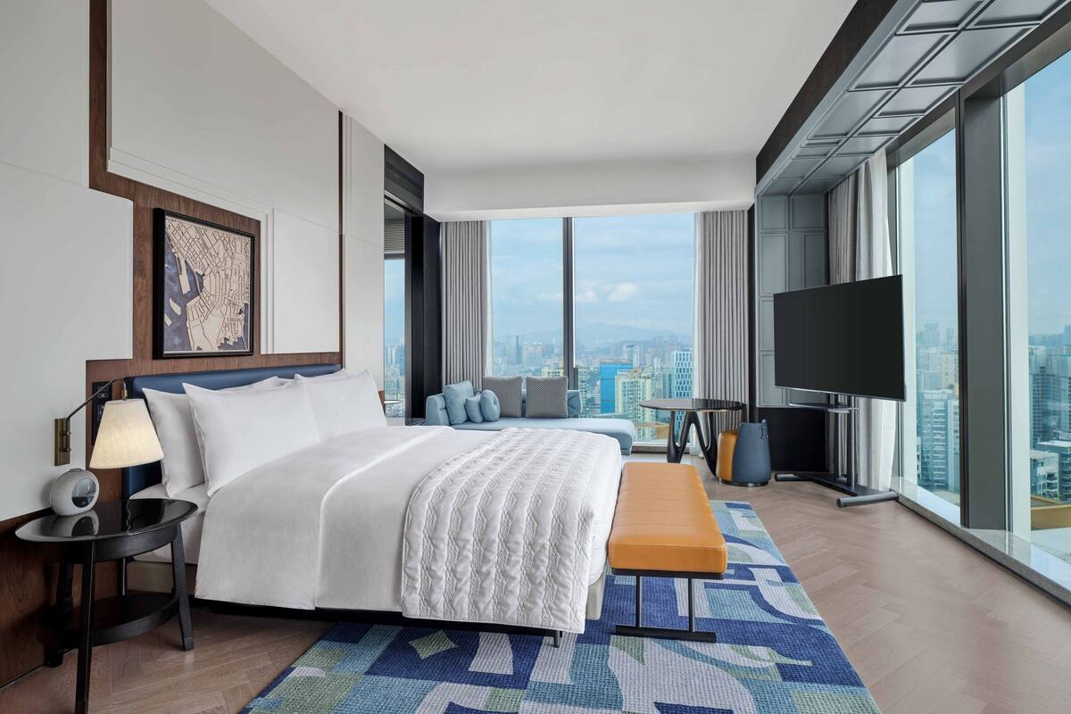 A modern hotel room with a large bed, a TV, a city view, and a small seating area.