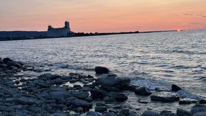Eine felsige Küste mit Sonnenuntergang im Hintergrund und einem Gebäude gegenüber dem Wasser.