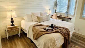 Quarto aconchegante com uma cama, duas mesas de cabeceira com abajures, um criado-mudo e uma janela com persianas, oferecendo vista para árvores.