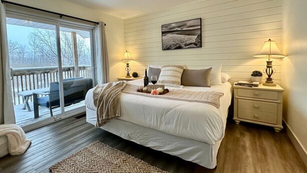 Quarto com uma cama grande, mesa de cabeceira com abajur, janela com vista para o exterior e um deck de madeira lá fora.