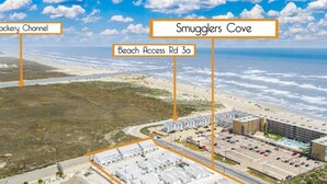 An aerial view of a coastal area with a parking lot, buildings, and a beach. - Paradise Palms - Walk to Beach! (Corpus Christi)