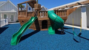 A play area with a slide and a swing set, surrounded by wooden play structures and a blue rubberized floor. - Paradise Palms - Walk to Beach! (Corpus Christi)