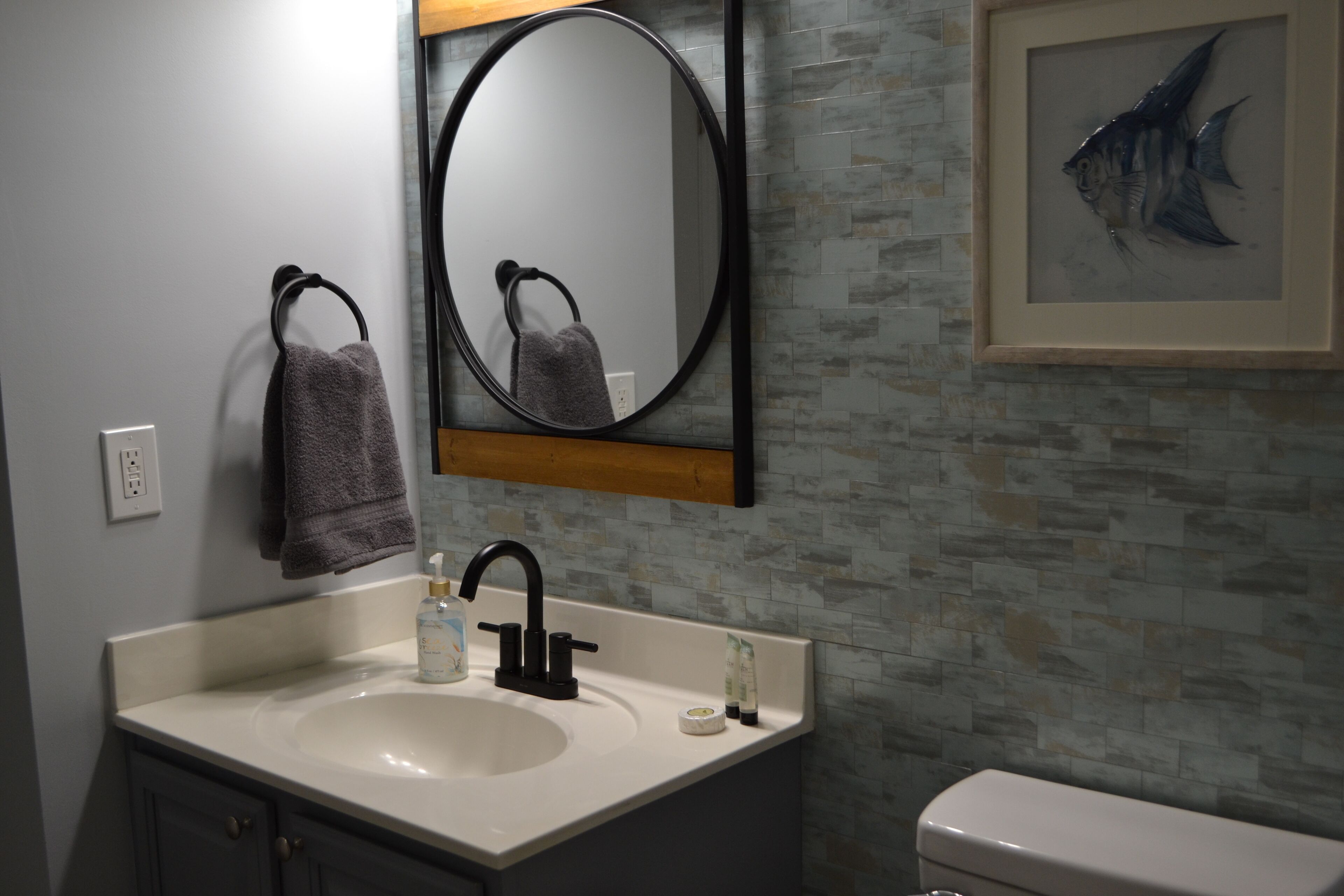 Un baño con un lavamanos blanco, grifo negro y un espejo encima. Se observa una obra de arte con peces en un marco en la pared y una toalla colgada en un perchero.