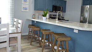 Eine moderne Küche mit blauen Schränken, einem Edelstahl-Kühlschrank, einer Mikrowelle und einer Marmorarbeitsplatte. Holzstühle und ein Esstisch mit Stühlen sind ebenfalls vorhanden.