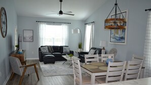 Ein modernes Wohnzimmer mit einer Couch, einem Esstisch und Stühlen. Es gibt einen Deckenventilator, eine Lüster und ein großes Fenster mit Vorhängen.
