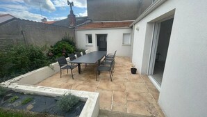 A patio with a table and chairs, a small garden, and a white building with a tiled roof. - House 10/12pers town center 5mn from Puy Du Fou "AU COEUR DU BOURG (Chanverrie)