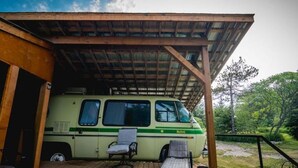 A green and white van parked under a wooden structure with a covered deck, surrounded by greenery. - Unique Glamping RV on 100 acres 1 hr from Toronto (Blackstock)