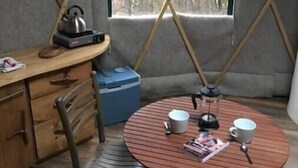 Une pièce chaleureuse avec une table en bois mise pour deux personnes, une bouilloire et une machine à café. Il y a une fenêtre donnant sur des arbres et une étagère contenant des ustensiles de cuisine.