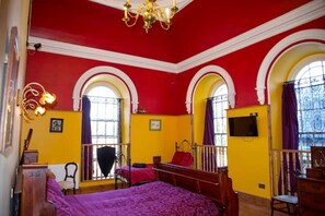 Ein Zimmer mit rotem und gelben Farbschema, zwei Betten, einer Kristallleuchtre und Bogenfenstern.